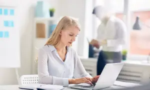 Kobieta pracuje na laptopie w nowoczesnym biurze. W tle mężczyzna czyta dokumenty. Nowoczesne biuro z office 2021.