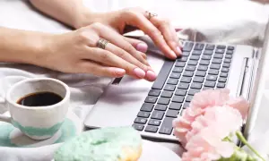 Dłonie z pierścionkami piszą na laptopie, obok kawa, pączki i kwiaty. Twarda spacja na klawiaturze czeka na naciśnięcie.