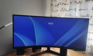 Zakrzywiony monitor 16:9 z niebieską tapetą i zegarem 09:43. Obok klawiatura i lampka.