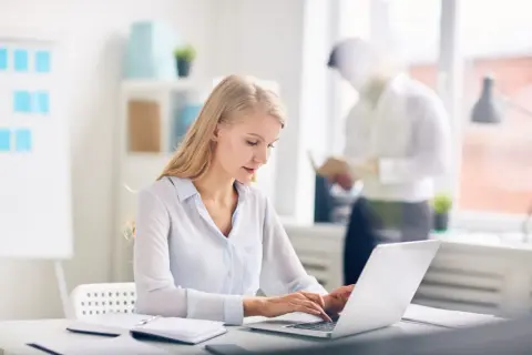 Kobieta pracuje na laptopie w nowoczesnym biurze. W tle mężczyzna czyta dokumenty. Nowoczesne biuro z office 2021.