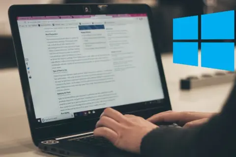 Osoba pisze na laptopie, na ekranie widać artykuł o malowaniu dachu. W tle widnieje niebieskie logo Windows.