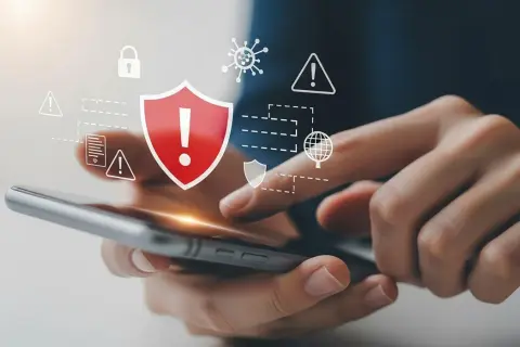 Dłonie trzymające smartfon, nad którym wyświetlają się ikony symbolizujące bezpieczeństwo i ochronę przed zagrożeniami, w tym czerwiona tarcza z wykrzyknikiem. To wizualizacja skutecznego **antywirusa na telefon**.