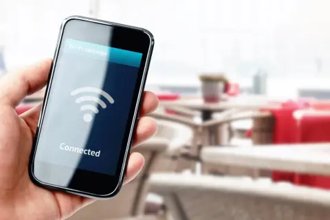 Dłoń trzyma smartfon z napisem "Connected" i ikoną Wi-Fi. W tle widać stoliki kawiarniane.