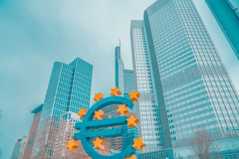 Duży, niebieski znak euro z żółtymi gwiazdami, na tle nowoczesnych wieżowców.