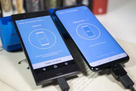Dwa telefony połączone kablem USB, jeden wyświetla komunikat "Gotowe", drugi "100%", pokazując jak przenieść dane z telefonu na telefon Samsung.
