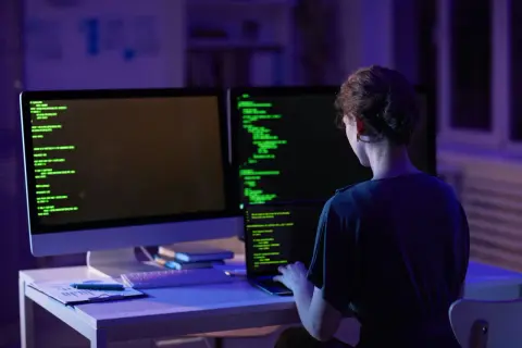 Kobieta programistka pracuje nocą nad kodem. Na ekranach widać zielony tekst, a w pomieszczeniu panuje niebiesko-fioletowe oświetlenie. To jej codzienny crud.