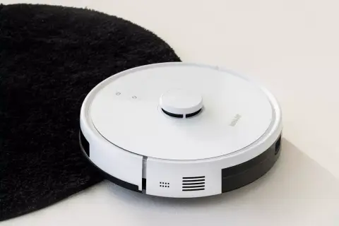 Biały robot sprzątający obok czarnego dywanika. Jego moc ssania odkurzacza poradzi sobie z każdym brudem.