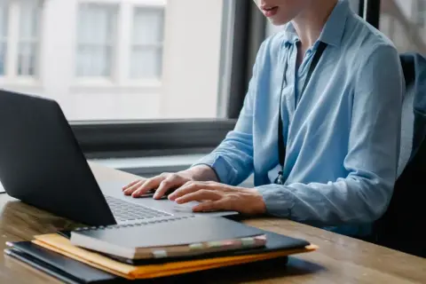 Osoba w niebieskiej koszuli pracuje przy laptopie, tworząc skrypty VBA. Na biurku leżą notatniki.