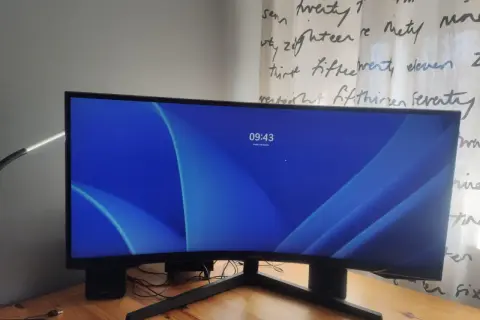 Zakrzywiony monitor 16:9 z niebieską tapetą i zegarem 09:43. Obok klawiatura i lampka.