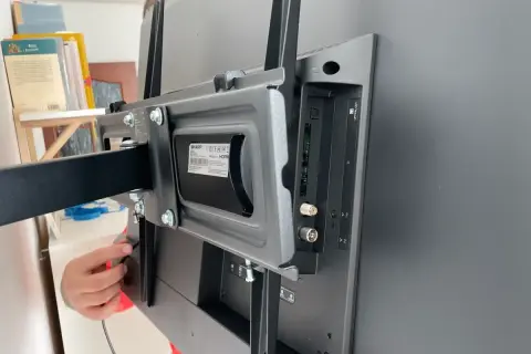 Montaż telewizora na ścianie: widok z tyłu na uchwyt i porty. Ręka podłącza kabel.