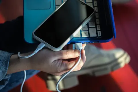 Dłoń podłącza kabel USB, tworząc łącze z telefonem leżącym na niebieskim laptopie.