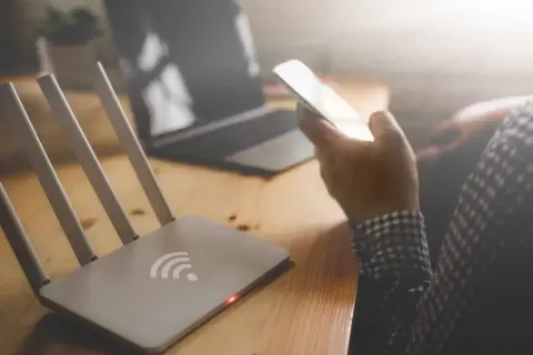 Router z czerwoną diodą i ręka trzymająca telefon. Brak internetu uniemożliwia pracę na laptopie.