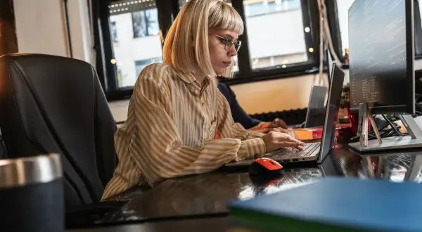 Kobieta w okularach i bluzce w paski pisze na laptopie, obok niej monitor z kodem. Wersaliki na ekranie i w tekście.
