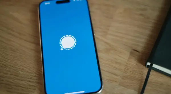 Telefon z niebieskim ekranem i ikoną komunikatora Signal. Obok leży czarny notatnik.