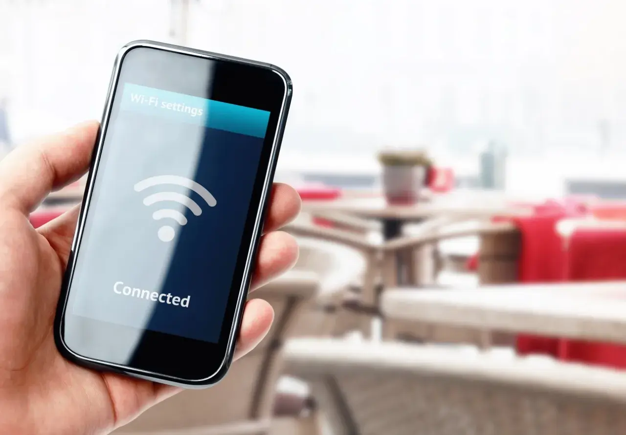 Dłoń trzyma smartfon z napisem "Connected" i ikoną Wi-Fi. W tle widać stoliki kawiarniane.