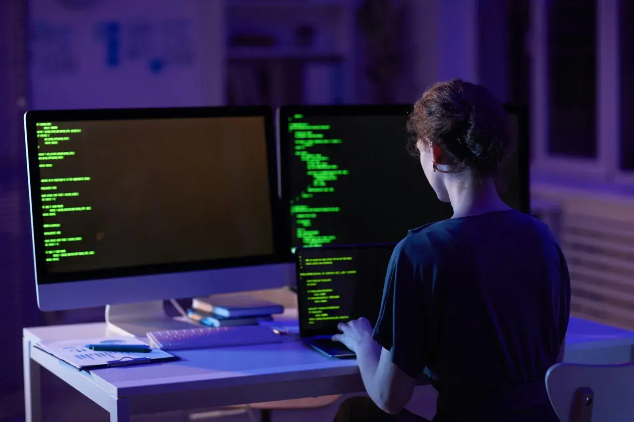 Kobieta programistka pracuje nocą nad kodem. Na ekranach widać zielony tekst, a w pomieszczeniu panuje niebiesko-fioletowe oświetlenie. To jej codzienny crud.