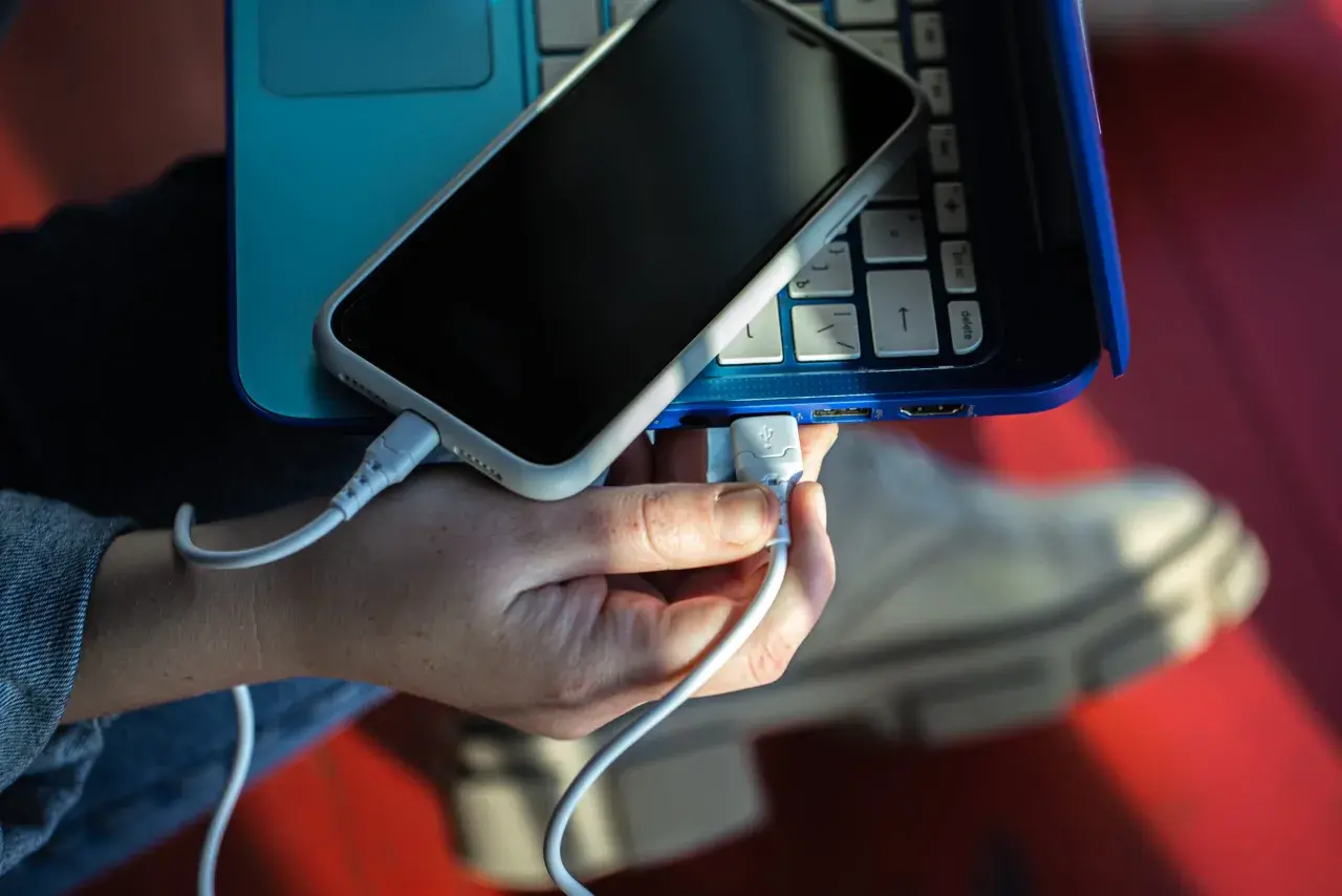 Dłoń podłącza kabel USB, tworząc łącze z telefonem leżącym na niebieskim laptopie.