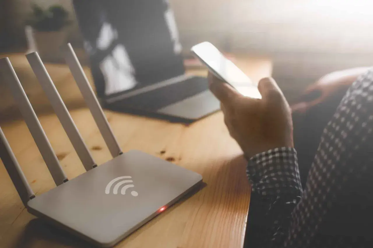Router z czerwoną diodą i ręka trzymająca telefon. Brak internetu uniemożliwia pracę na laptopie.