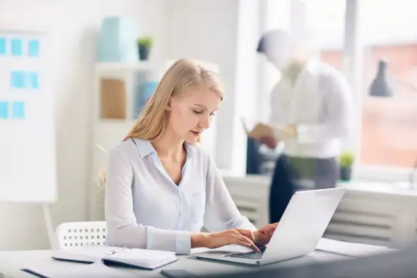 Kobieta pracuje na laptopie w nowoczesnym biurze. W tle mężczyzna czyta dokumenty. Nowoczesne biuro z office 2021.